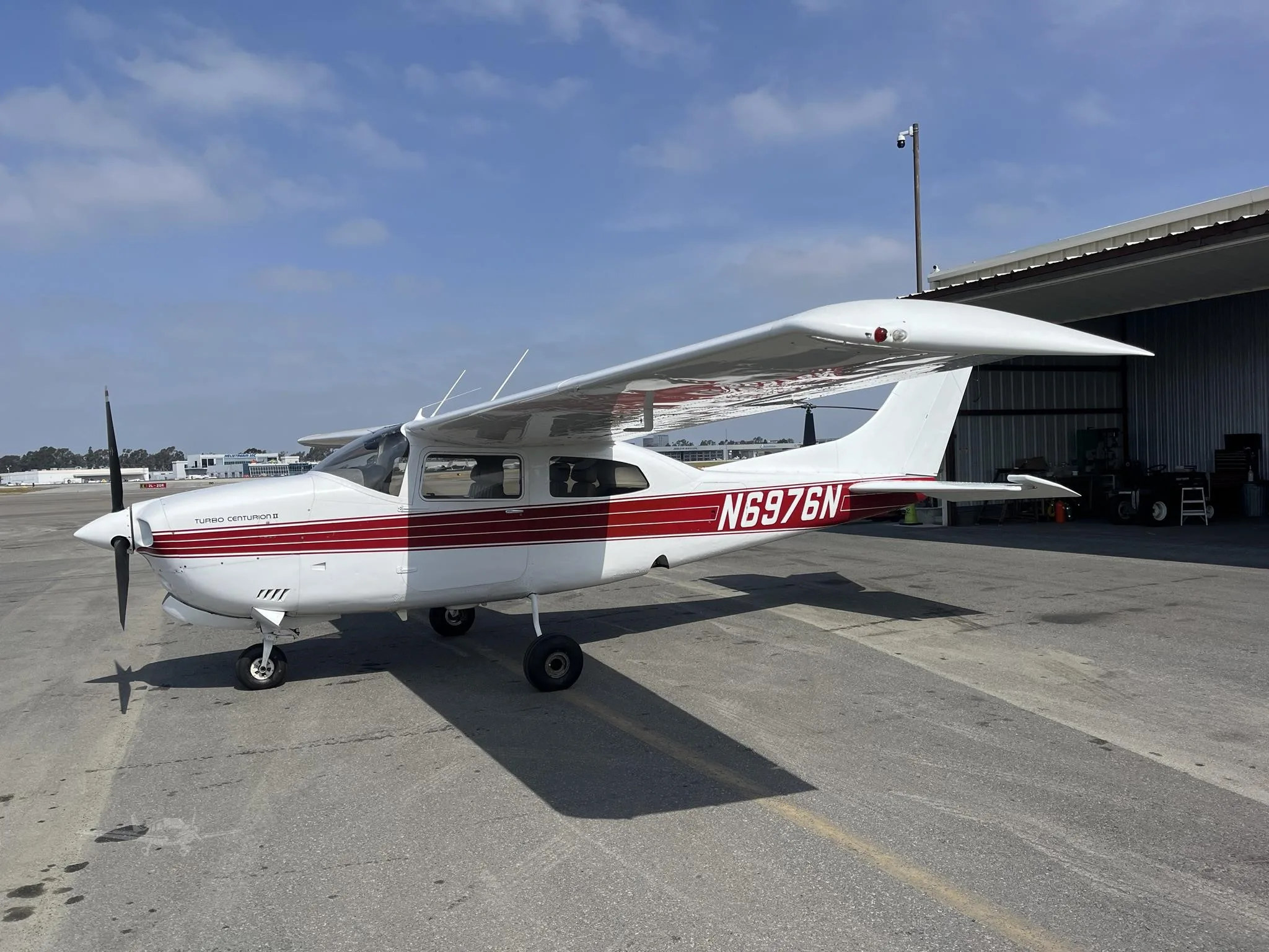 A white Cessna Turbo Centurion II with red stripes is parked on an airport apron with a hangar in the background.