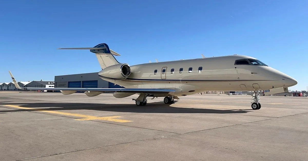 A beige and blue business jet, possibly a Learjet, parked on an airport tarmac next to a hangar under a clear sky.