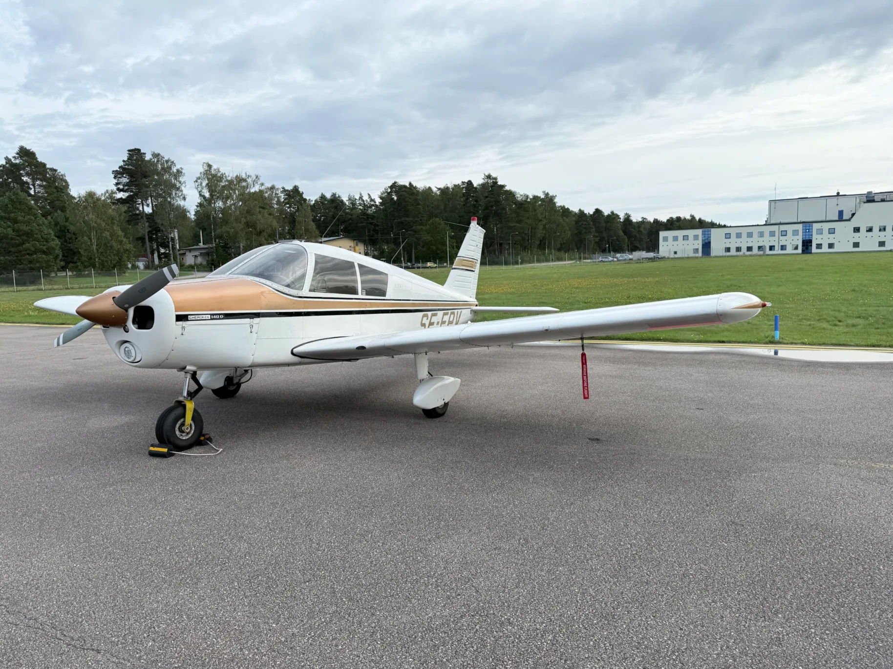 A white and gold Piper Cherokee 140D aircraft is parked on an asphalt apron with trees and a building in the background under a cloudy sky.