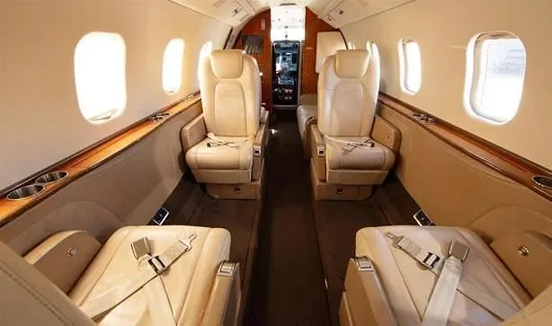 The luxurious interior cabin of a private jet, featuring beige leather seats and wood trim along the windows.