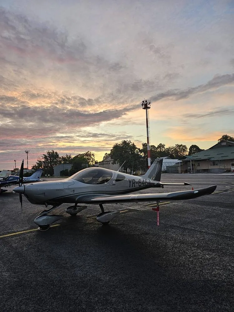 A silver and dark grey Pipistrel Virus SW aircraft (YR-6464) parked on an airfield at sunset.