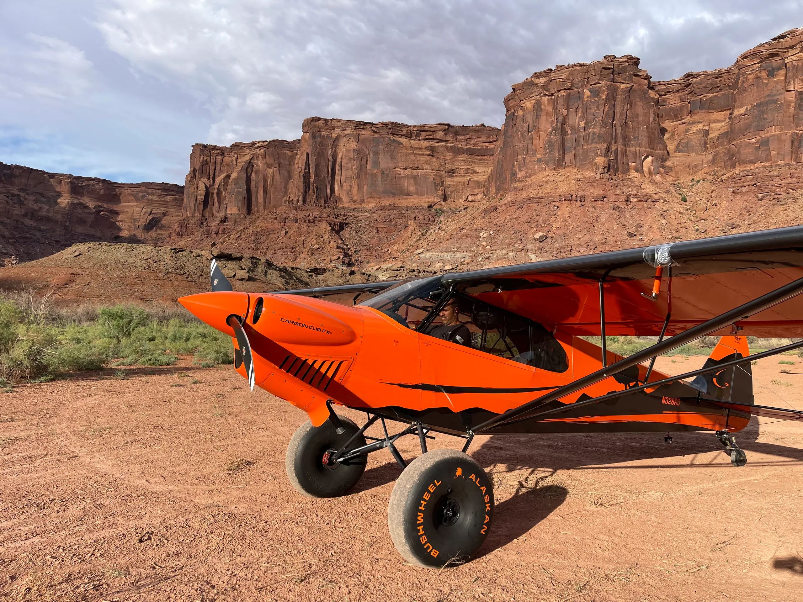 An orange and black Carbon Cub FX-3 aircraft with large bush wheels parked on red dirt in front of large rock formations.
