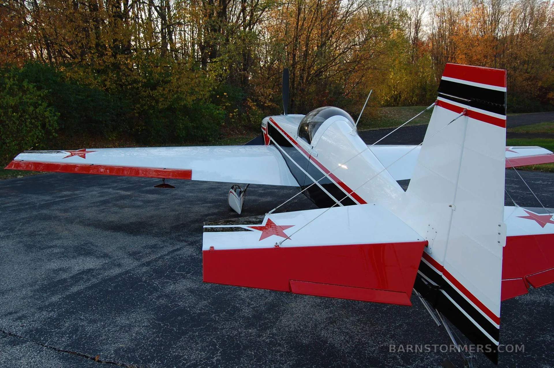 A white, black, and red Staudacher 300 aerobatic aircraft is parked on an asphalt surface with autumn trees in the background, viewed from the rear-left.