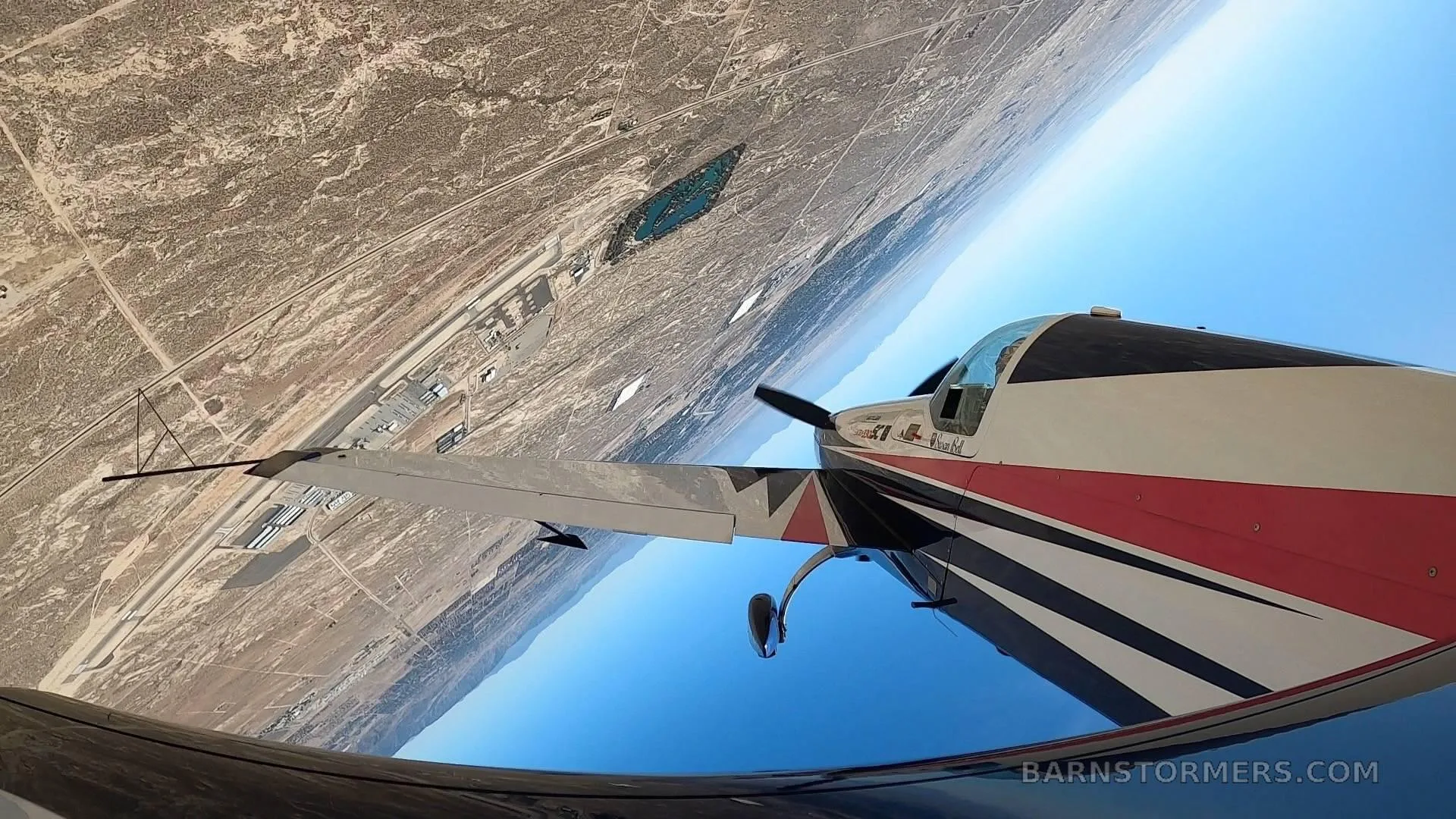 An Extra 330SC aircraft with red, white, and black stripes performs an inverted maneuver over a desert landscape.