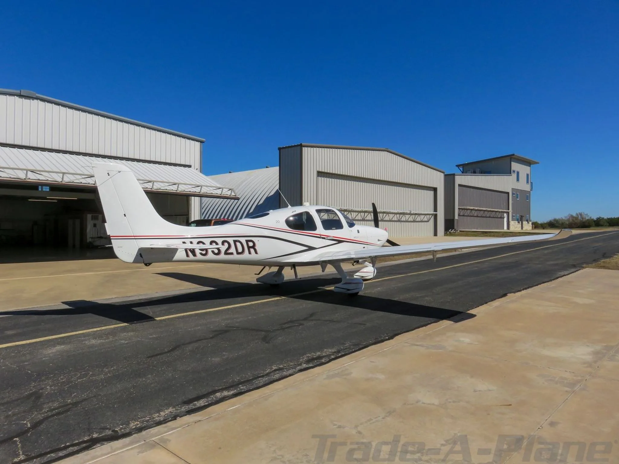 Three-quarter rear view of a white single-engine aircraft with red and black stripes, registration N932DR, parked on a tarmac near hangars.