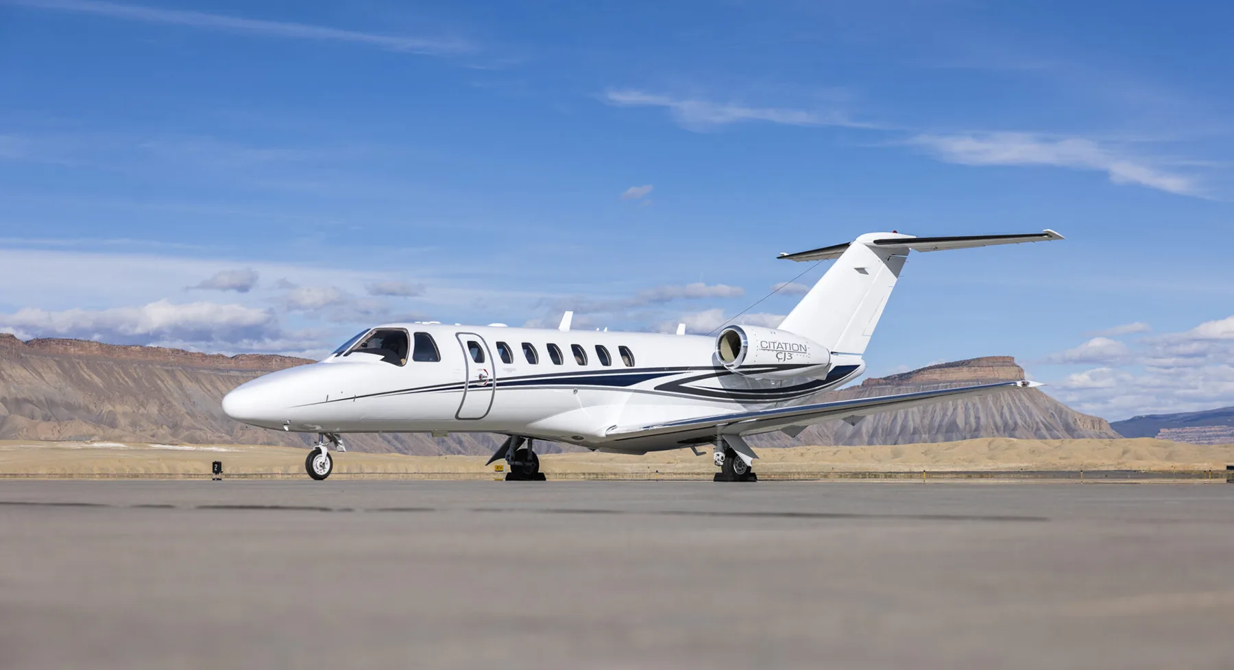 A white Citation CJ3 business jet with blue and black stripes parked on an airfield with desert mountains in the background.