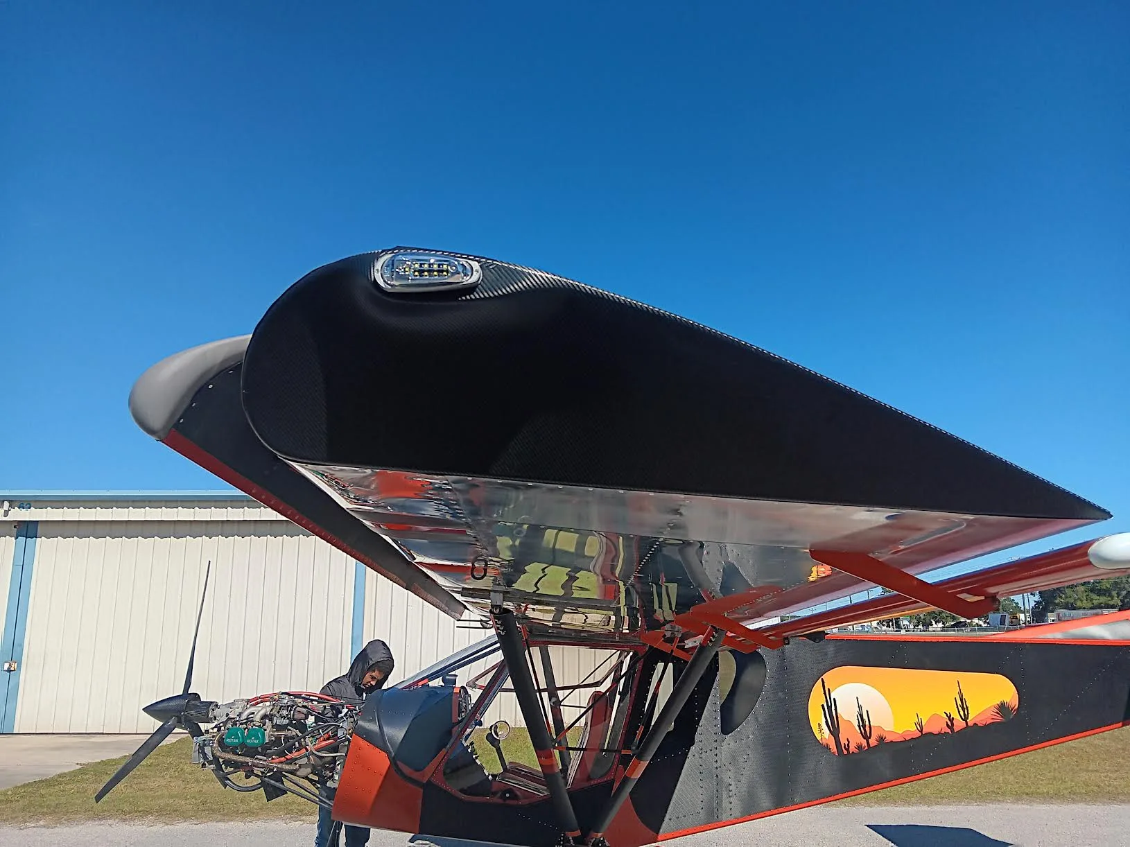 Close-up of the front and top of an orange and black ultralight aircraft with a desert sunset mural, parked outdoors.