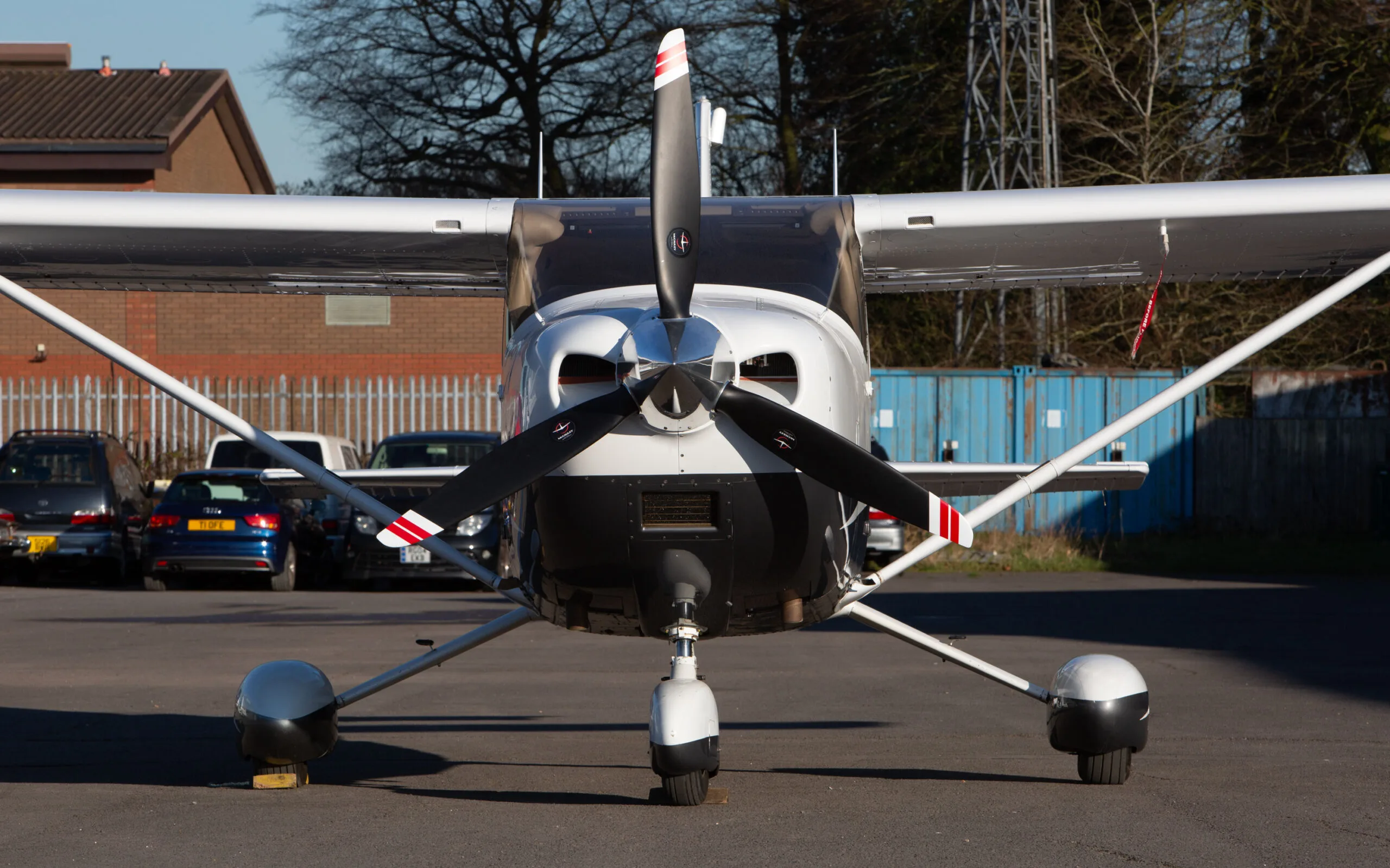 Front view of a white and black Cessna 182T (G-CYRL) parked on an airfield with buildings in the background.