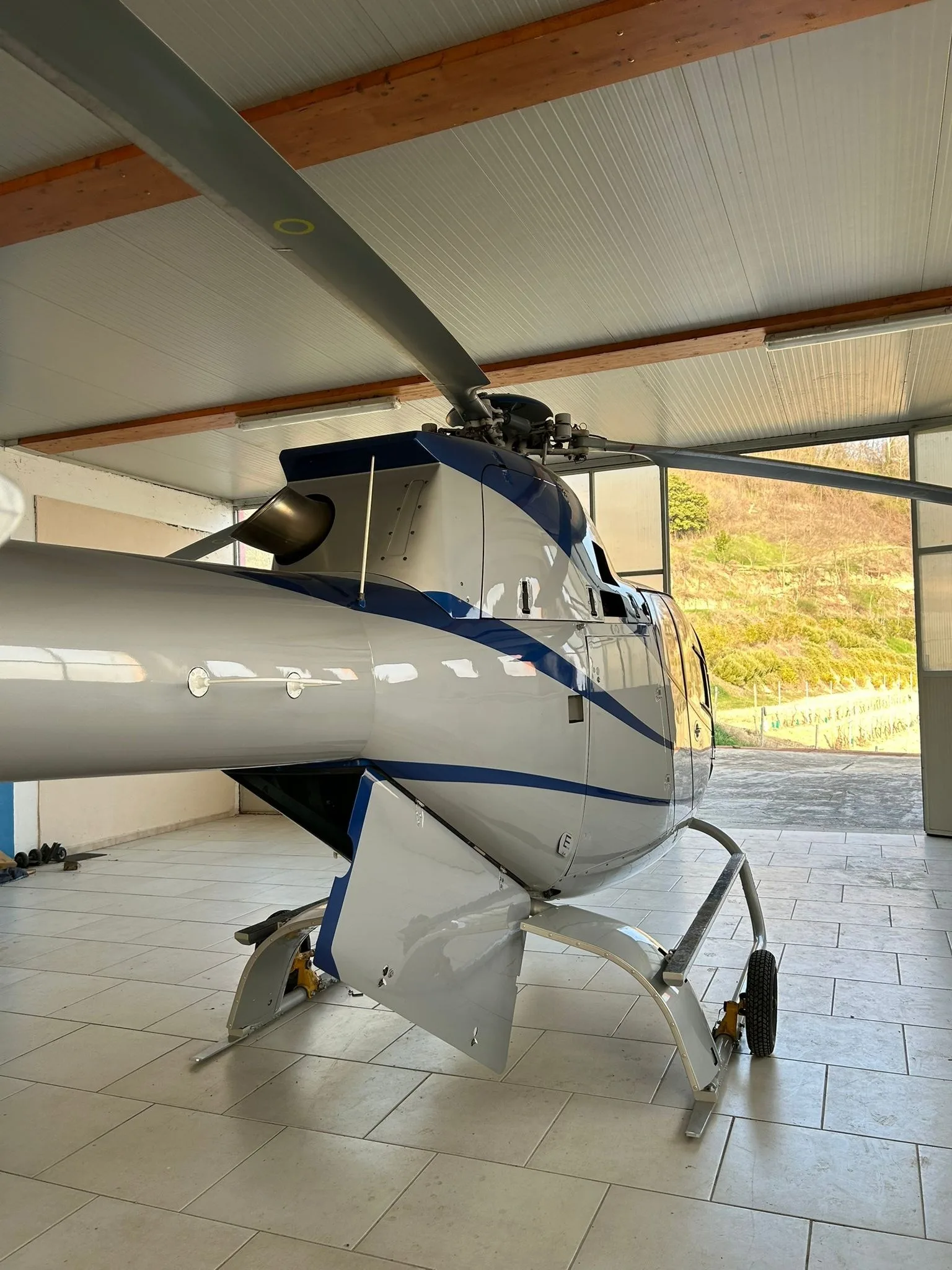 Rear-side view of an Airbus H120 Colibri helicopter in blue and silver livery inside a hangar, showing the tail boom and skid landing gear.