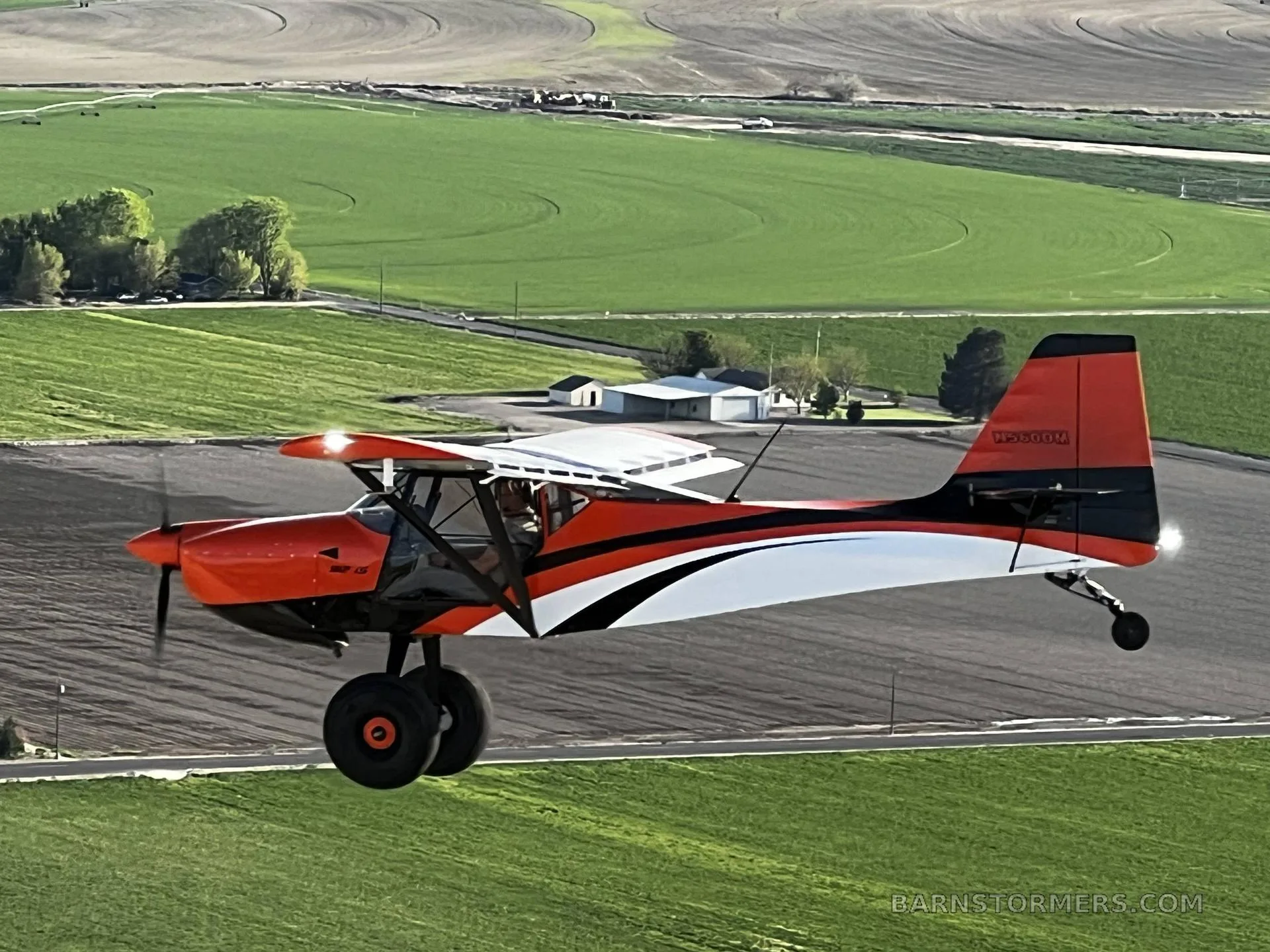 An orange and white Kitfox S7 Super Sport aircraft with large tires flying low over green agricultural fields.