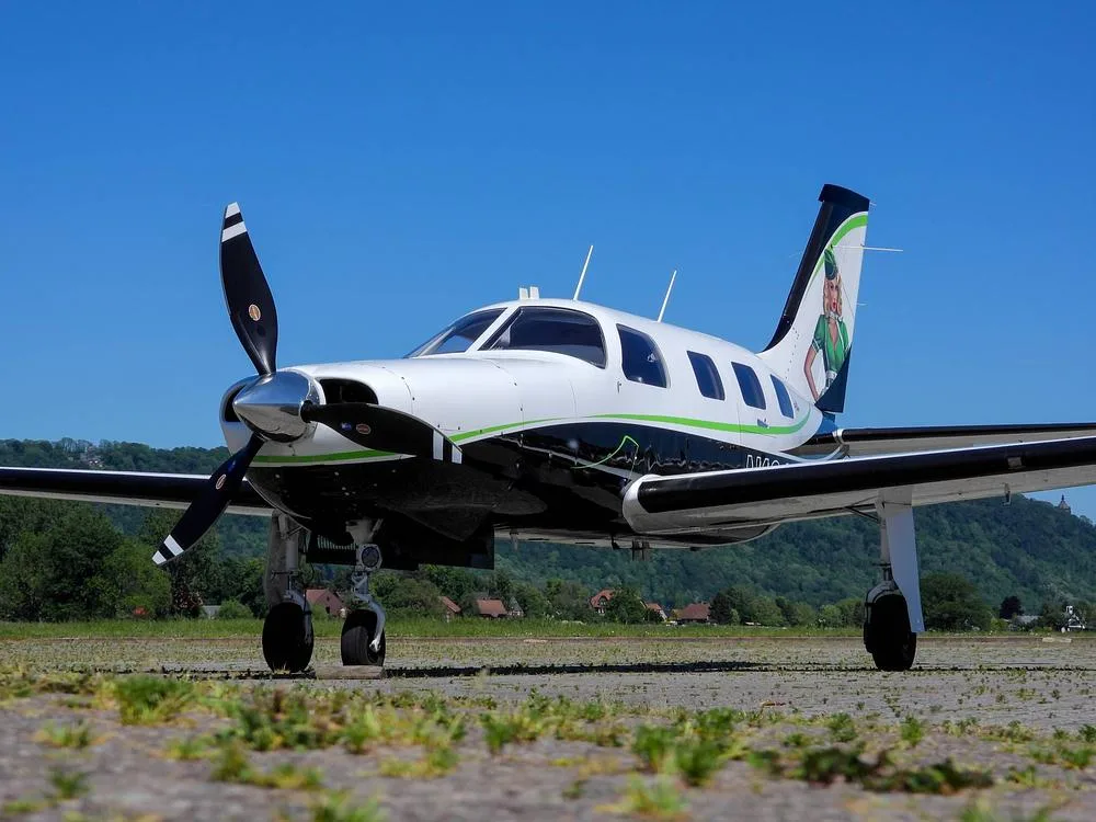 A low-angle front view of a white and black Piper M-Class aircraft with green accents and a pin-up girl on the tail, parked on a paved surface.