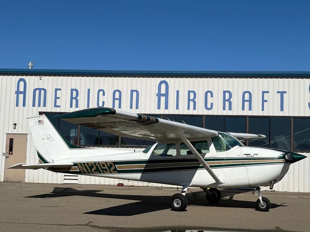 A white Cessna 172 with green and gold stripes parked on an airfield in front of a hangar.