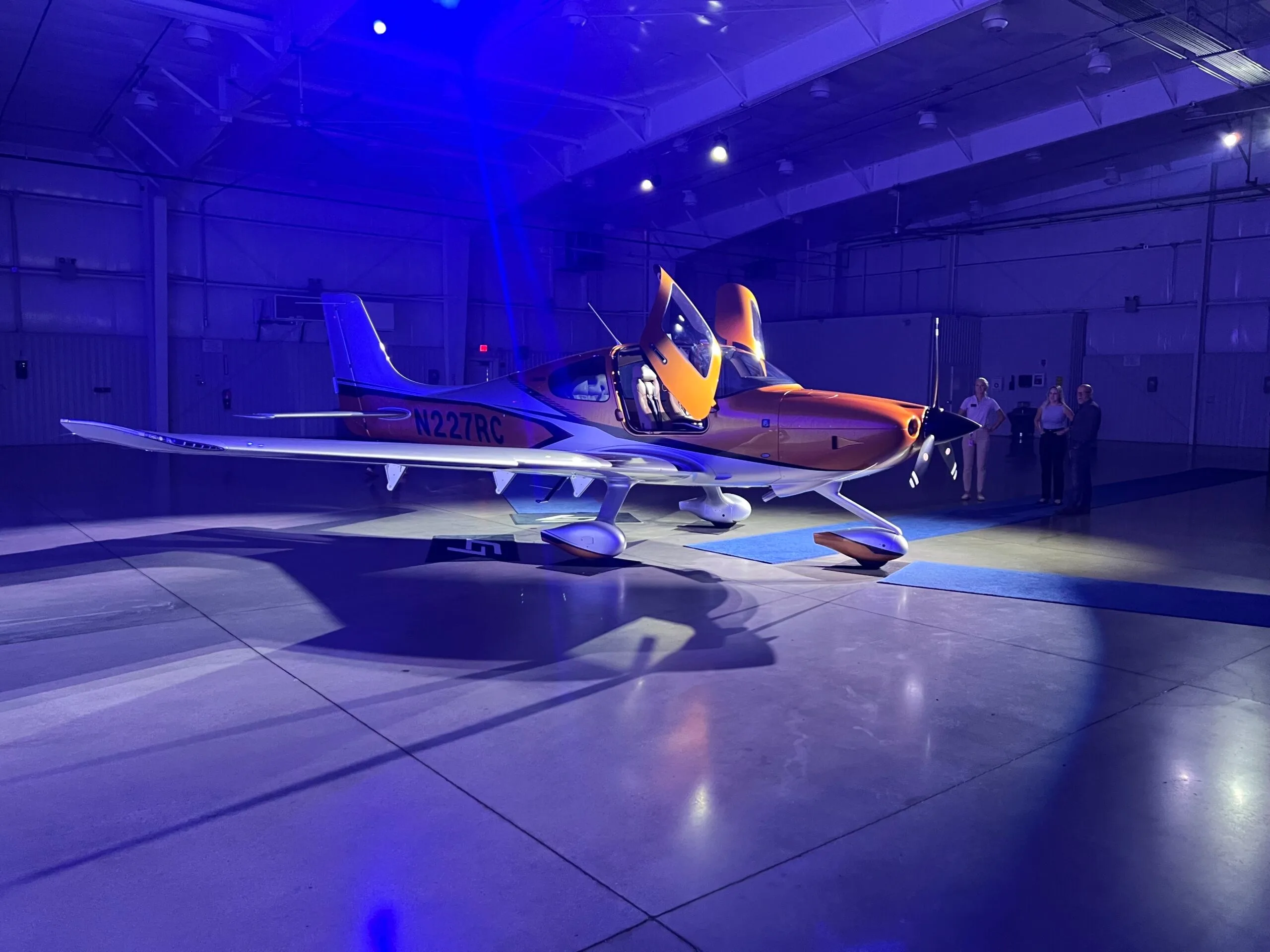 A Cirrus SR22 aircraft with orange and silver paint, doors open, displayed in a hangar with blue lighting.