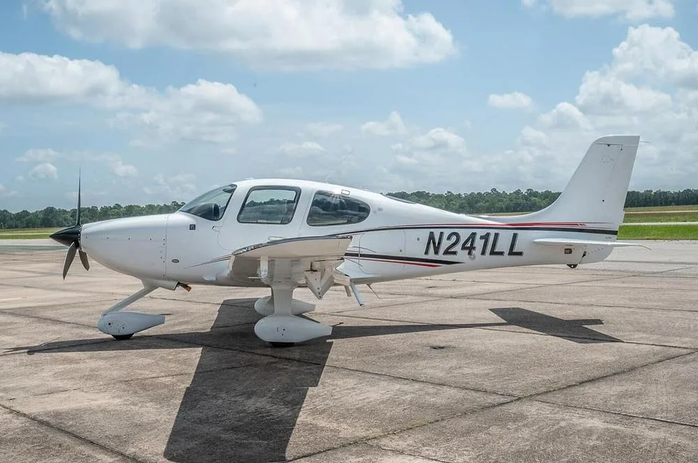 A white Cirrus SR22 aircraft with black and red stripes, registration N241LL, parked on an airfield under a partly cloudy sky.