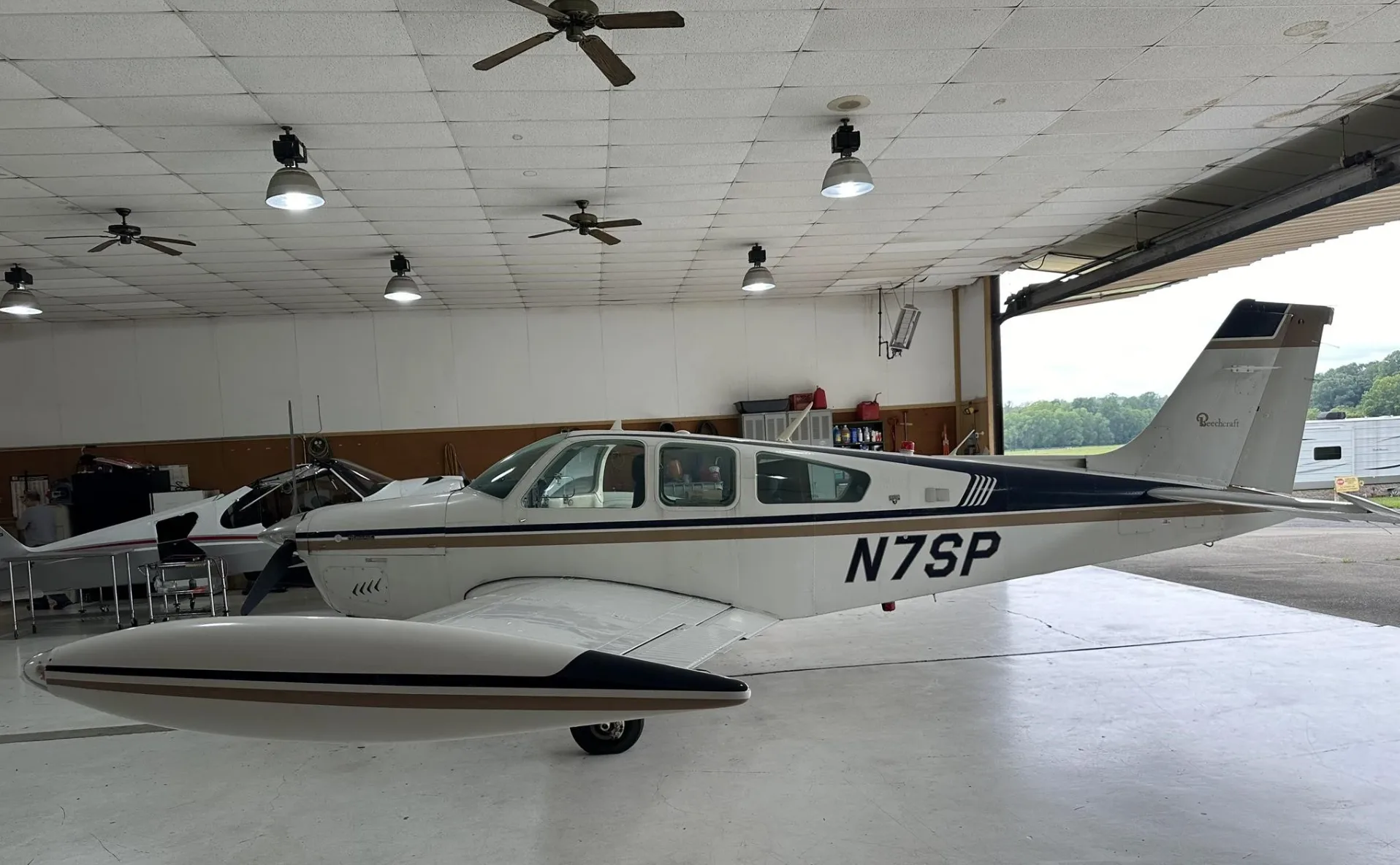 A white and gold Beechcraft Bonanza N7SP with tip tanks, parked in a hangar, viewed from the side.