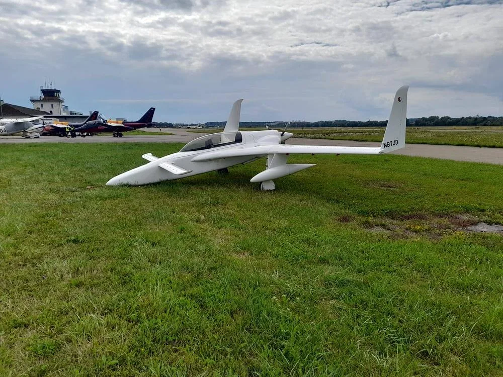 A white, single-engine aircraft with registration N97JD sits on grass next to a tarmac with a control tower in the background under a cloudy sky.
