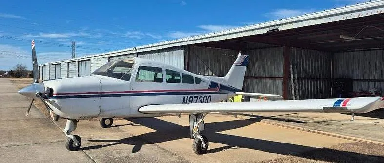 A white single-engine propeller aircraft with blue and red stripes, registration N9730Q, parked on an tarmac next to an open hangar under a blue sky.