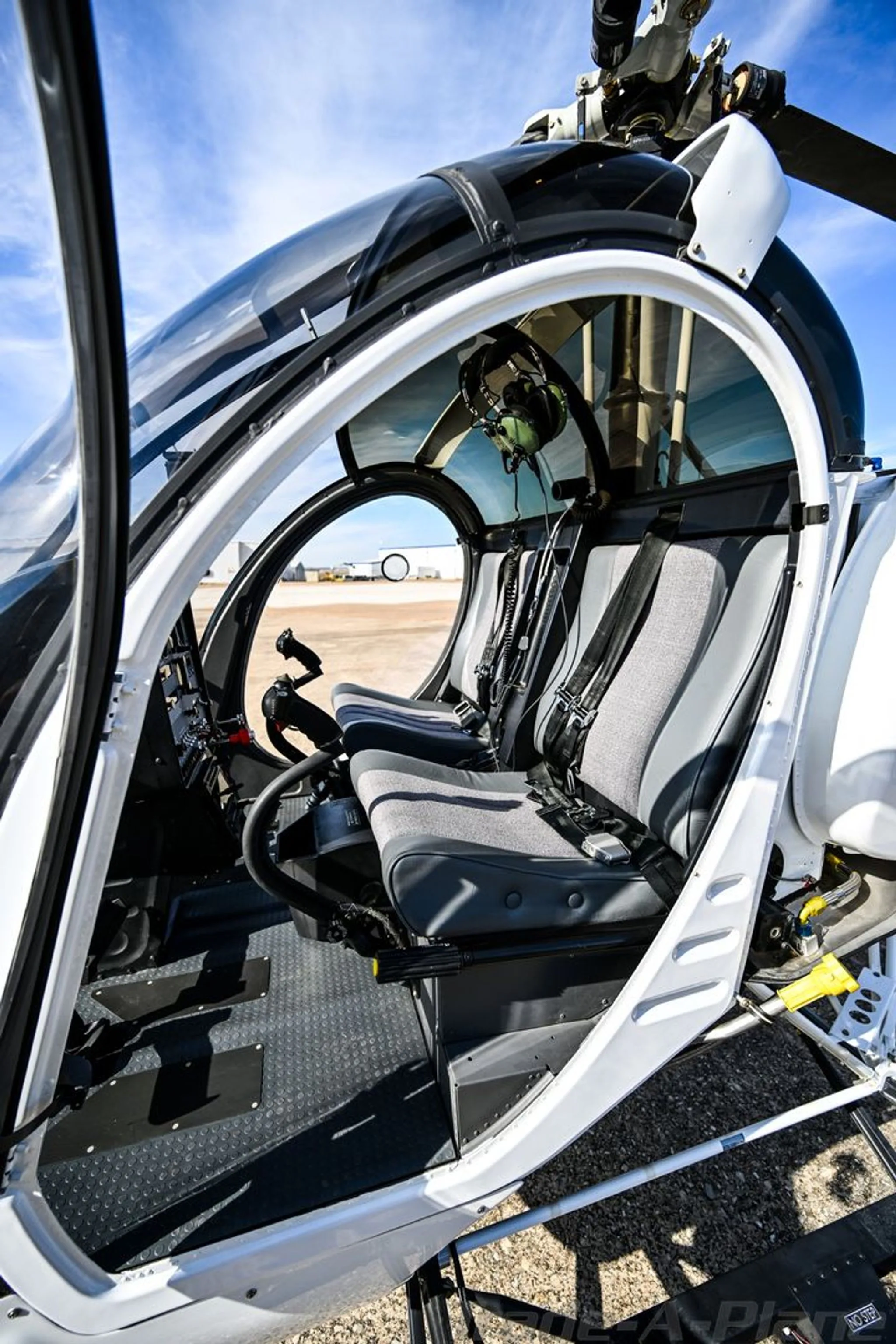 The interior of a white Schweizer S300CBi helicopter shows two grey and black seats with harnesses, with the door open.