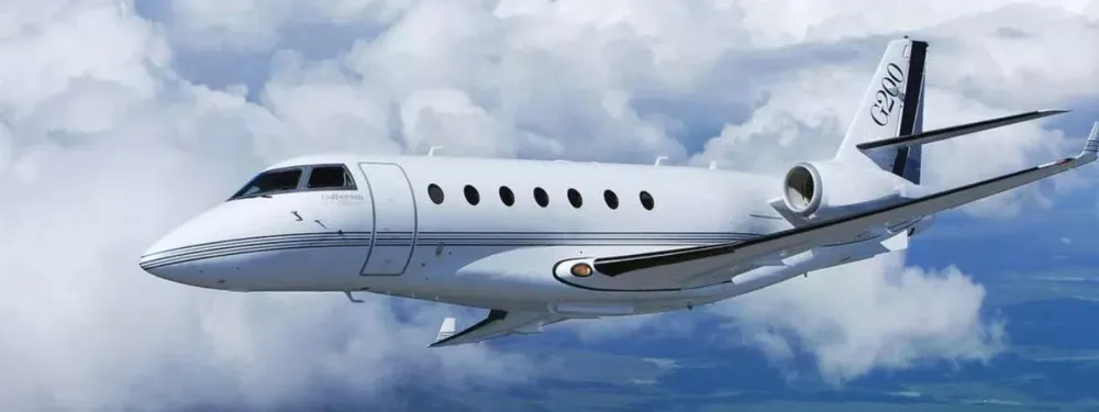 A white Gulfstream G200 business jet with black stripes on the tail flies through a cloudy sky.