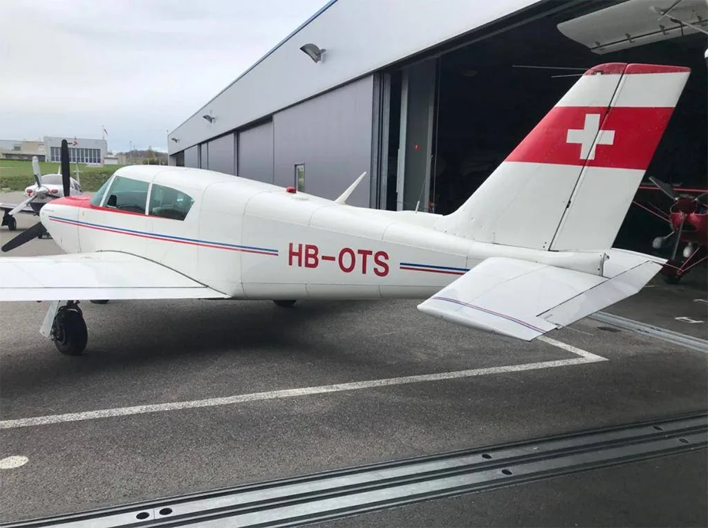 A white single-engine aircraft with red and blue stripes and a Swiss flag on the tail, parked outside a hangar.