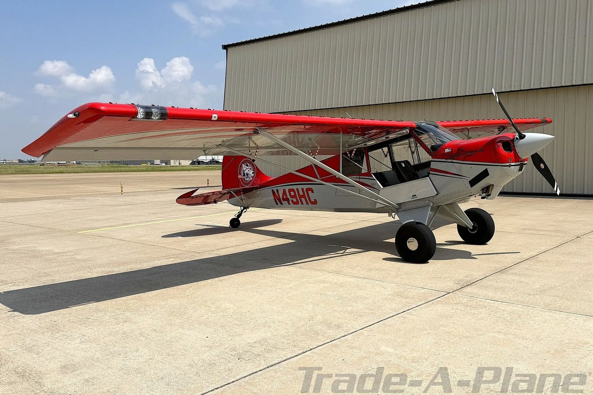 A red and white Aviat Husky A-1C aircraft, registration N49HC, parked on an airfield tarmac next to a hangar.