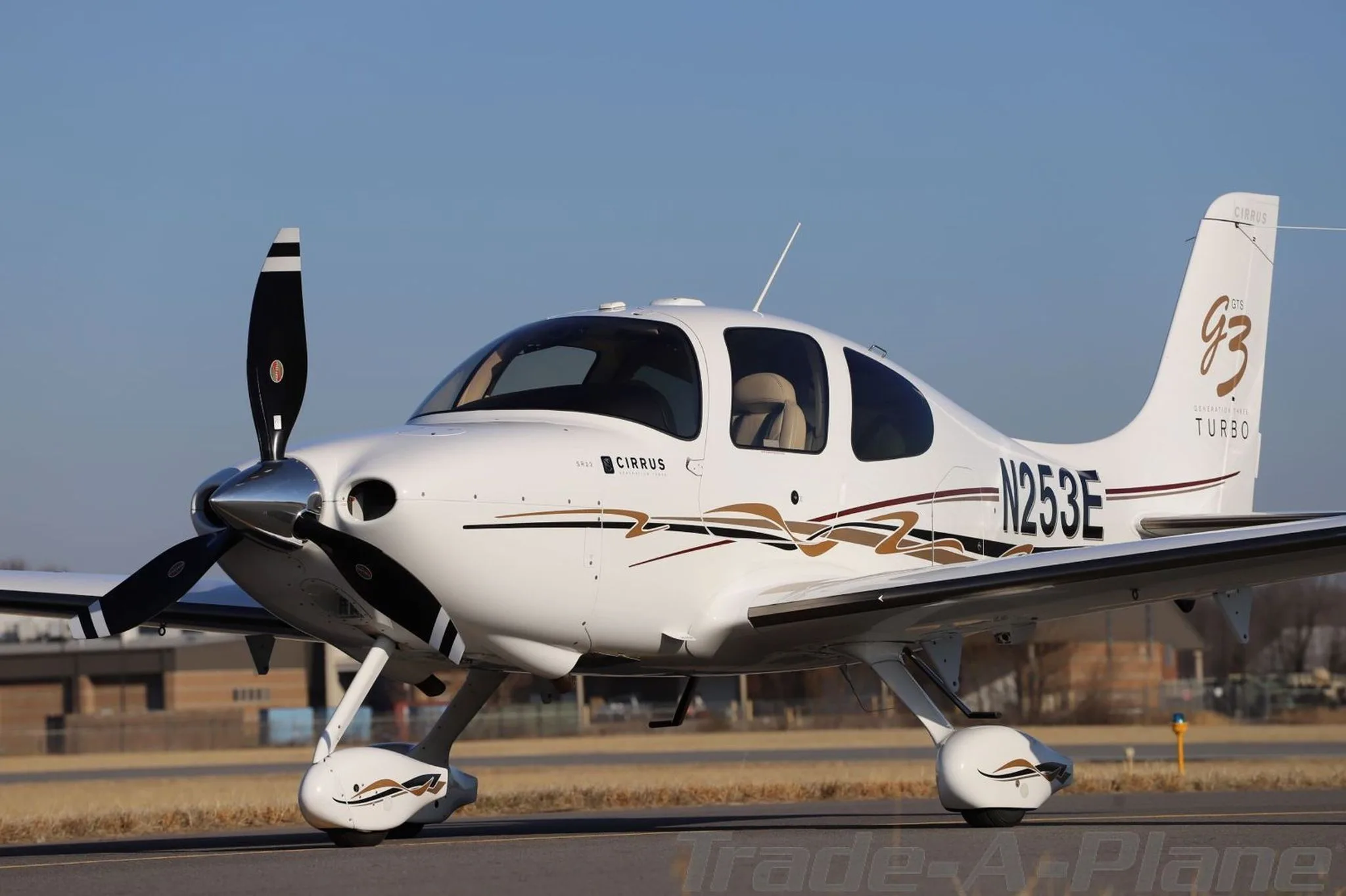 Side-front view of a white Cirrus SR22T aircraft (N253E) with gold and brown stripe livery, parked on an airfield.