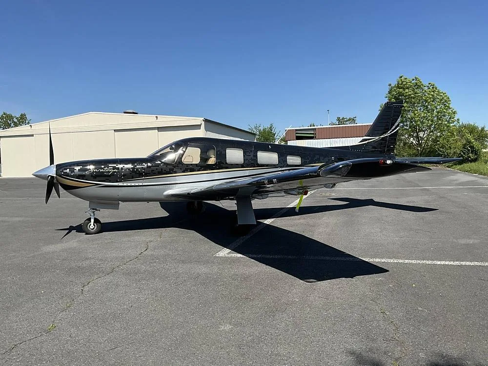 A black and silver Piper Saratoga aircraft with gold accents parked on a tarmac next to a hangar.