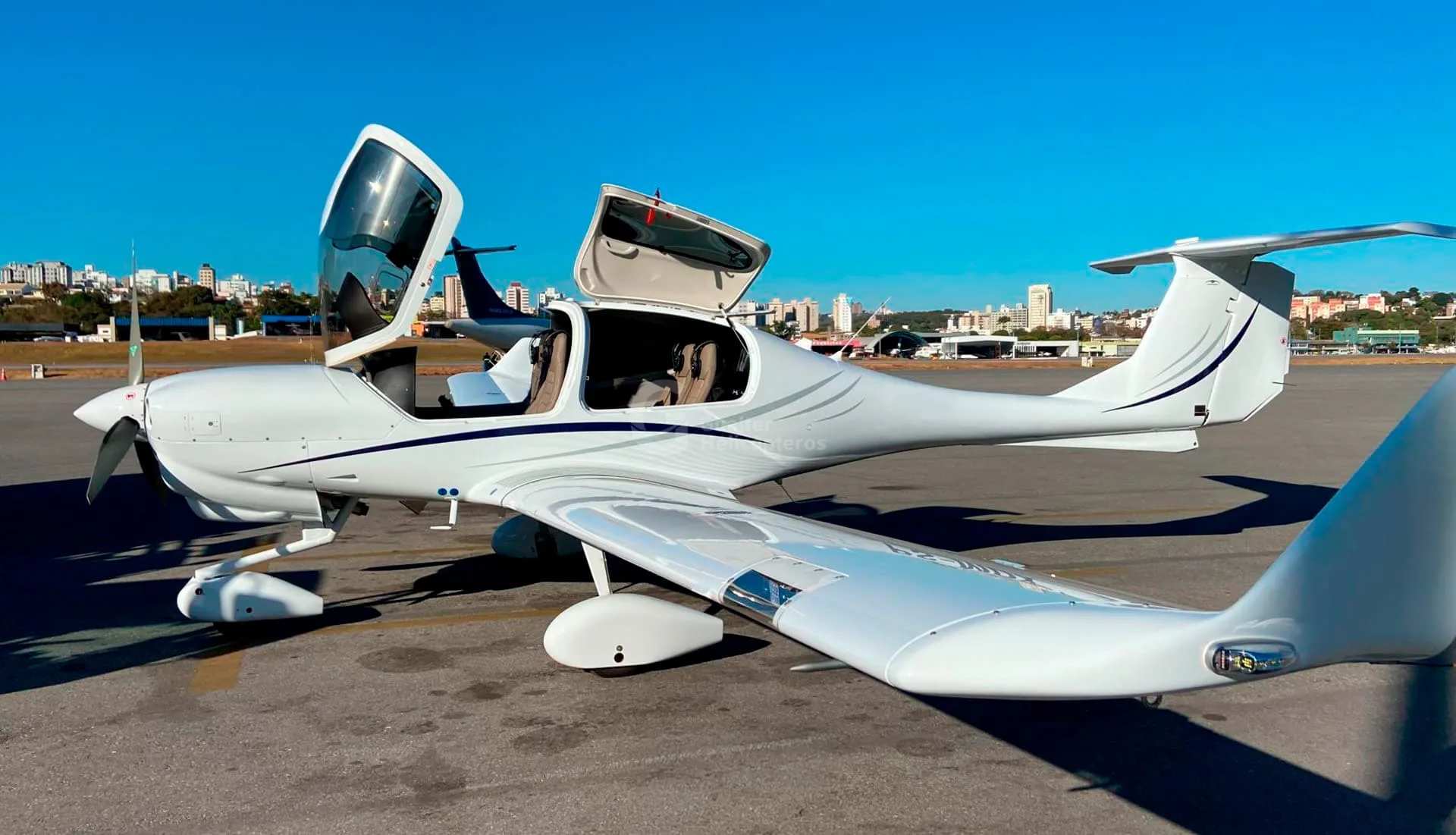 A white Diamond DA40 aircraft with open canopy doors parked on an airfield with a city skyline in the background.