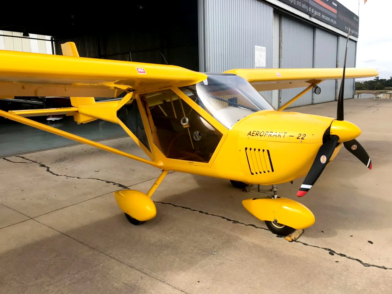 A three-quarter front view of a yellow Aeroprakt-22 aircraft parked outside a hangar.