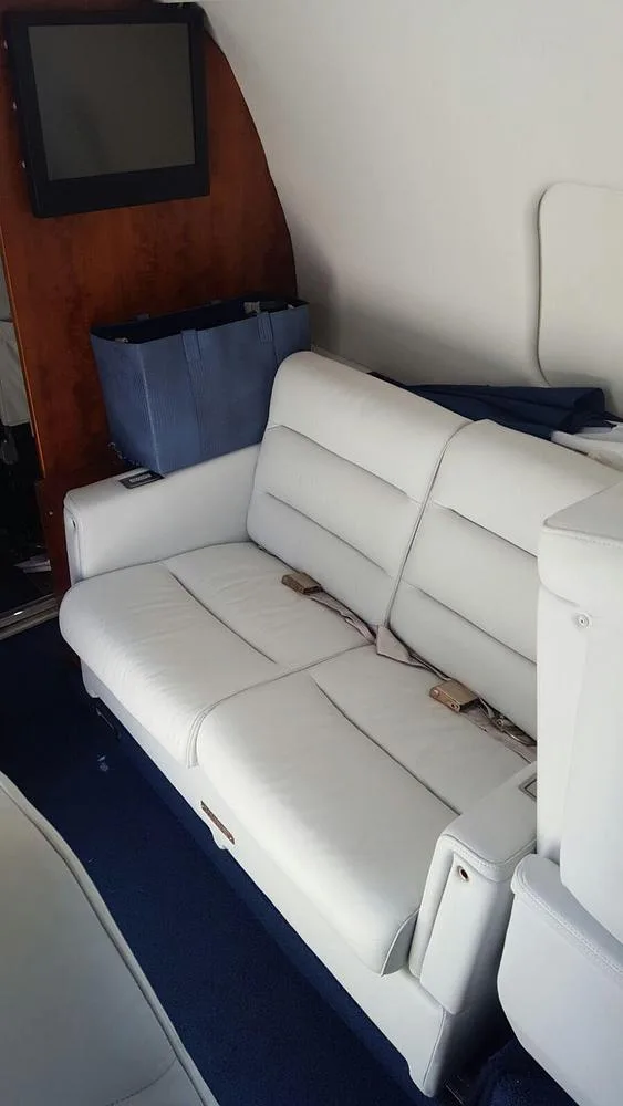 A two-person light grey leather couch seat in the private jet's cabin, next to wood paneling.