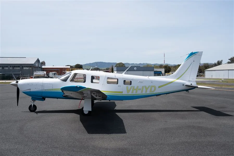 A white and blue Piper Seneca II HP aircraft (VH-IYD) parked on an asphalt tarmac, viewed from the side.