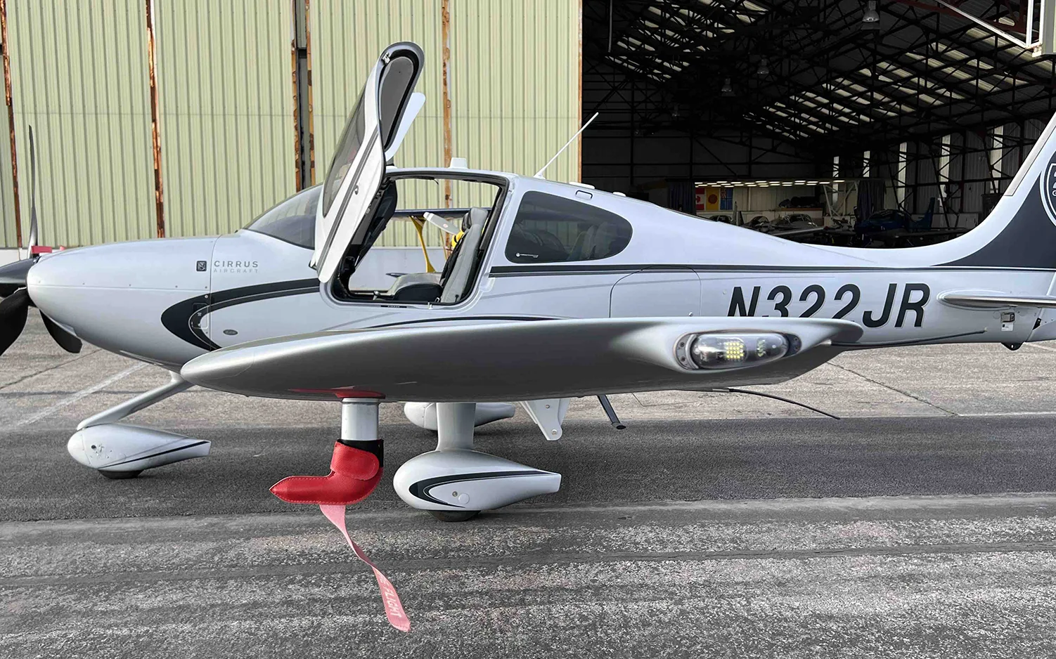 A silver Cirrus SR22 aircraft with registration N322JR and a red "REMOVE BEFORE FLIGHT" tag is parked next to a hangar.