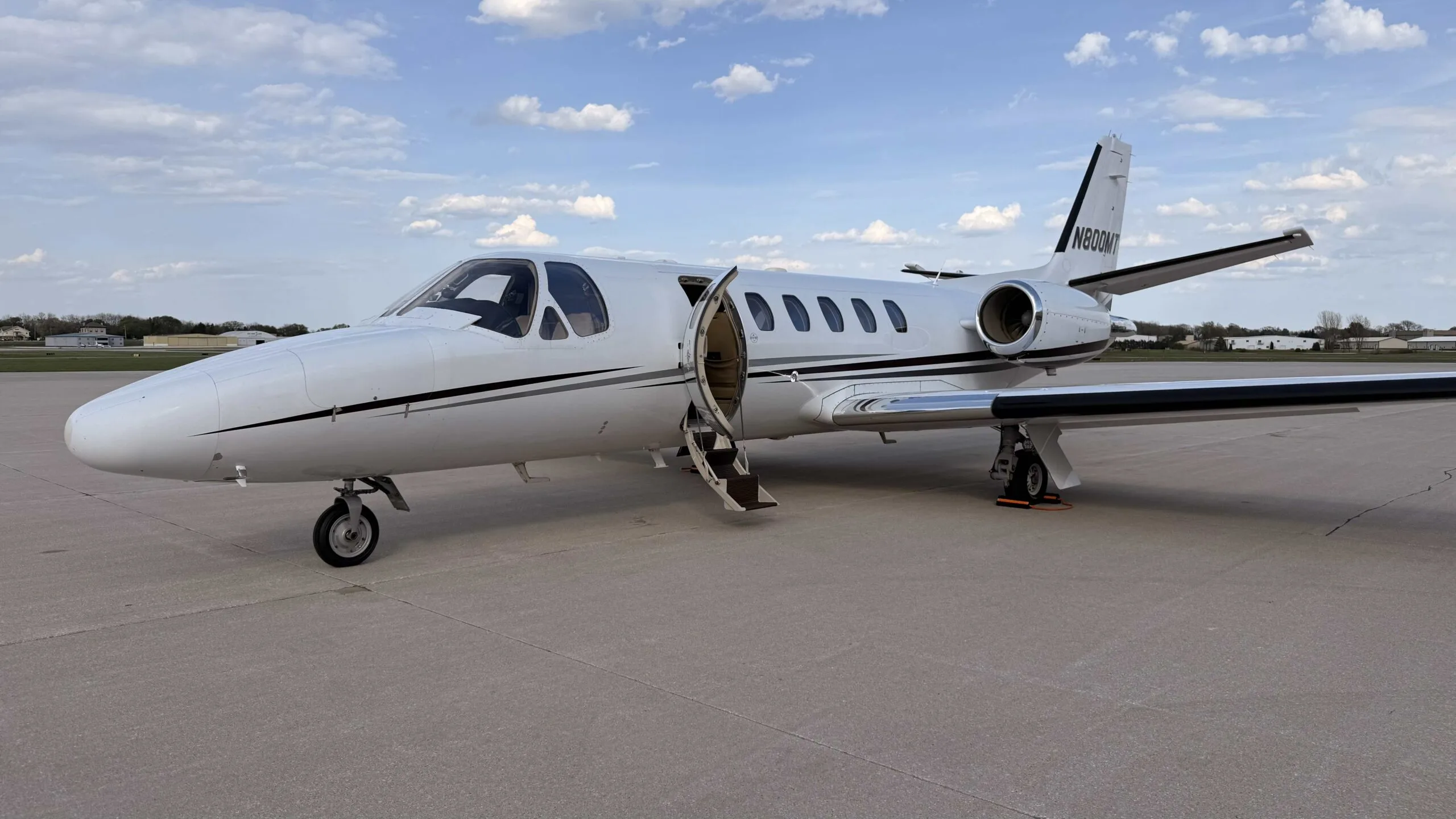 A white business jet with black stripes is parked on a tarmac with its main cabin door open under a partly cloudy sky.
