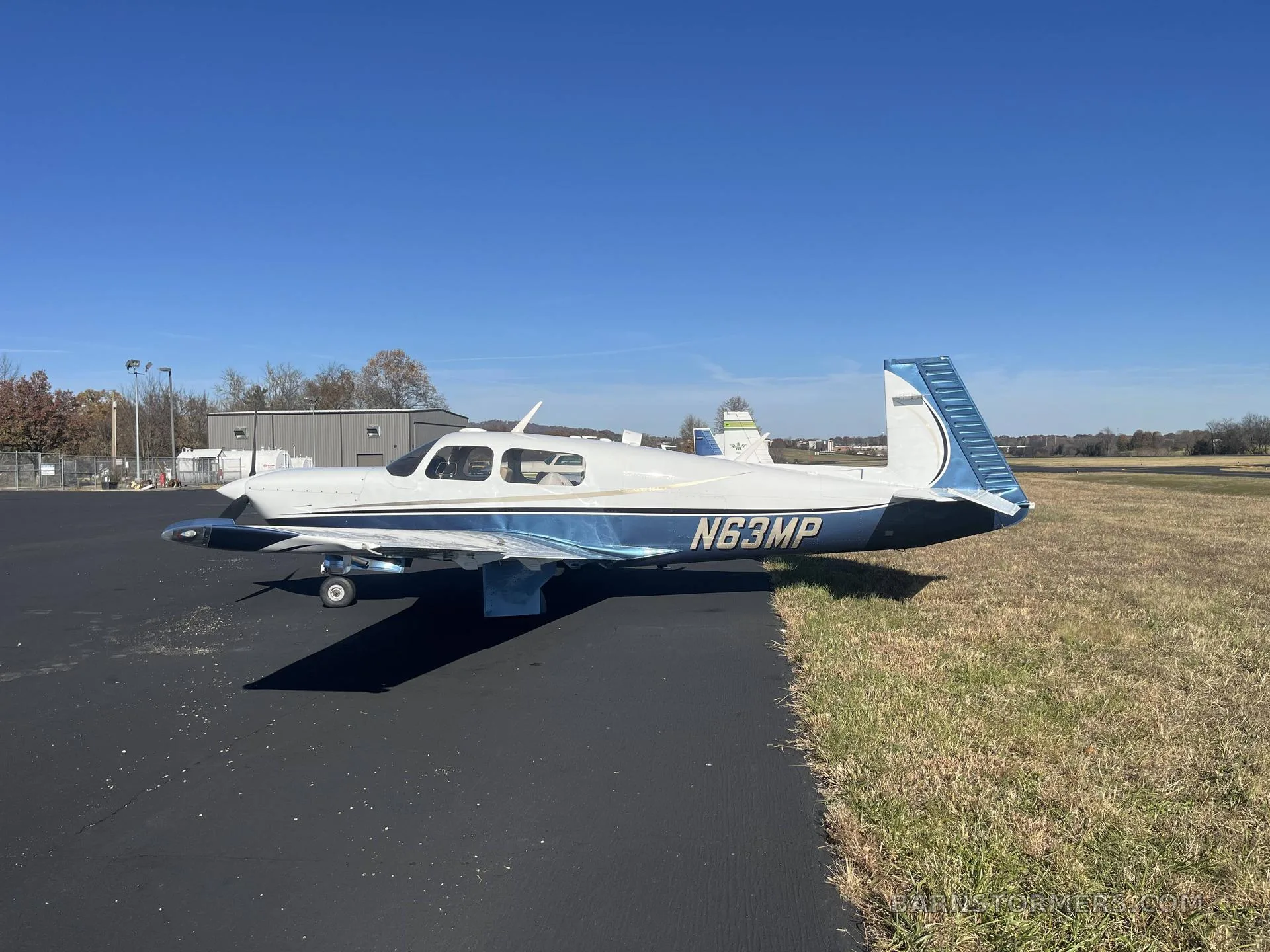 A white and blue single-engine propeller aircraft (N63MP) parked on an asphalt ramp with grass on the right, under a clear blue sky.