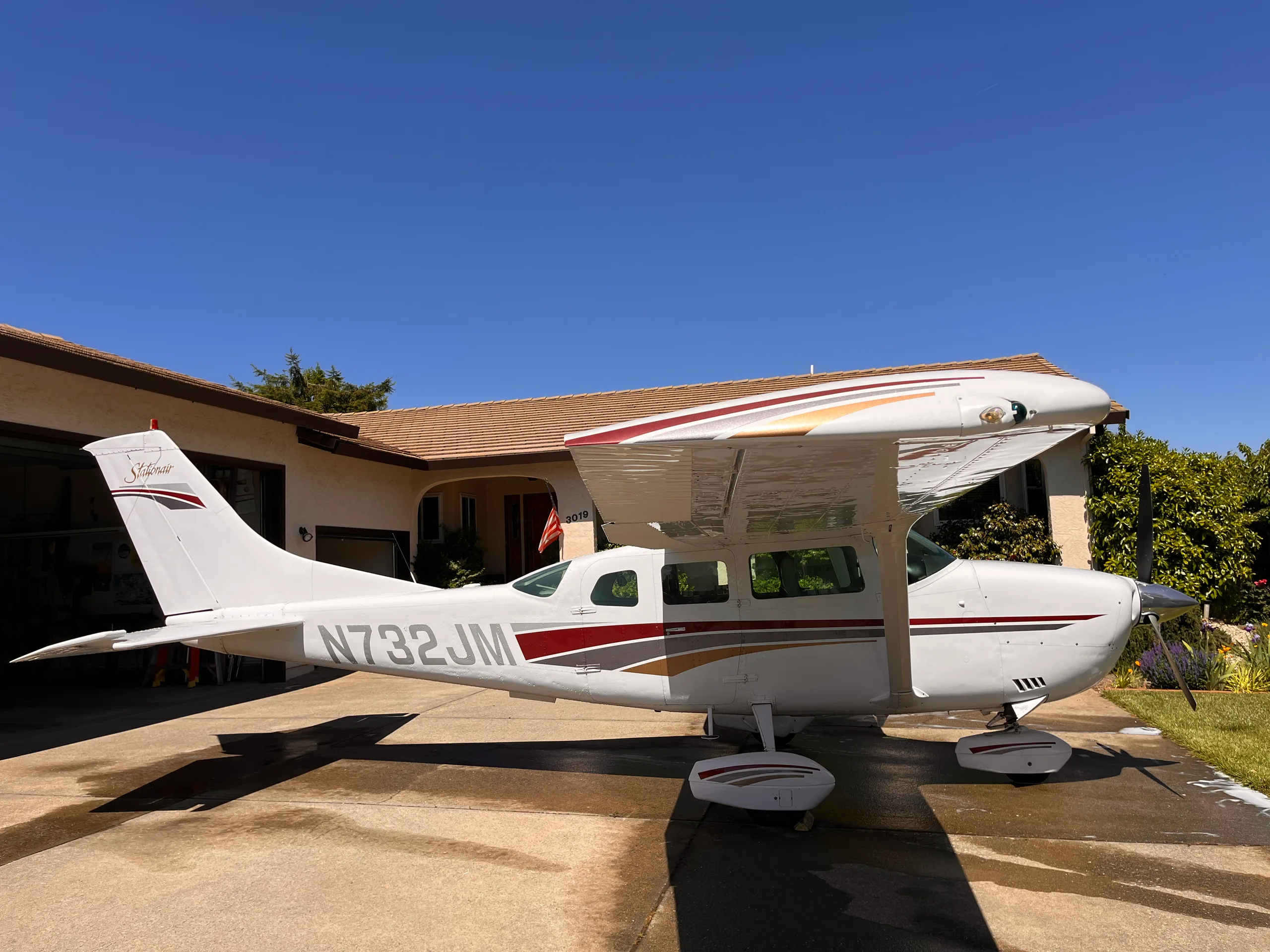 A white Cessna Stationair (N732JM) with red, grey, and gold stripes is parked on a concrete driveway in front of a house on a sunny day.