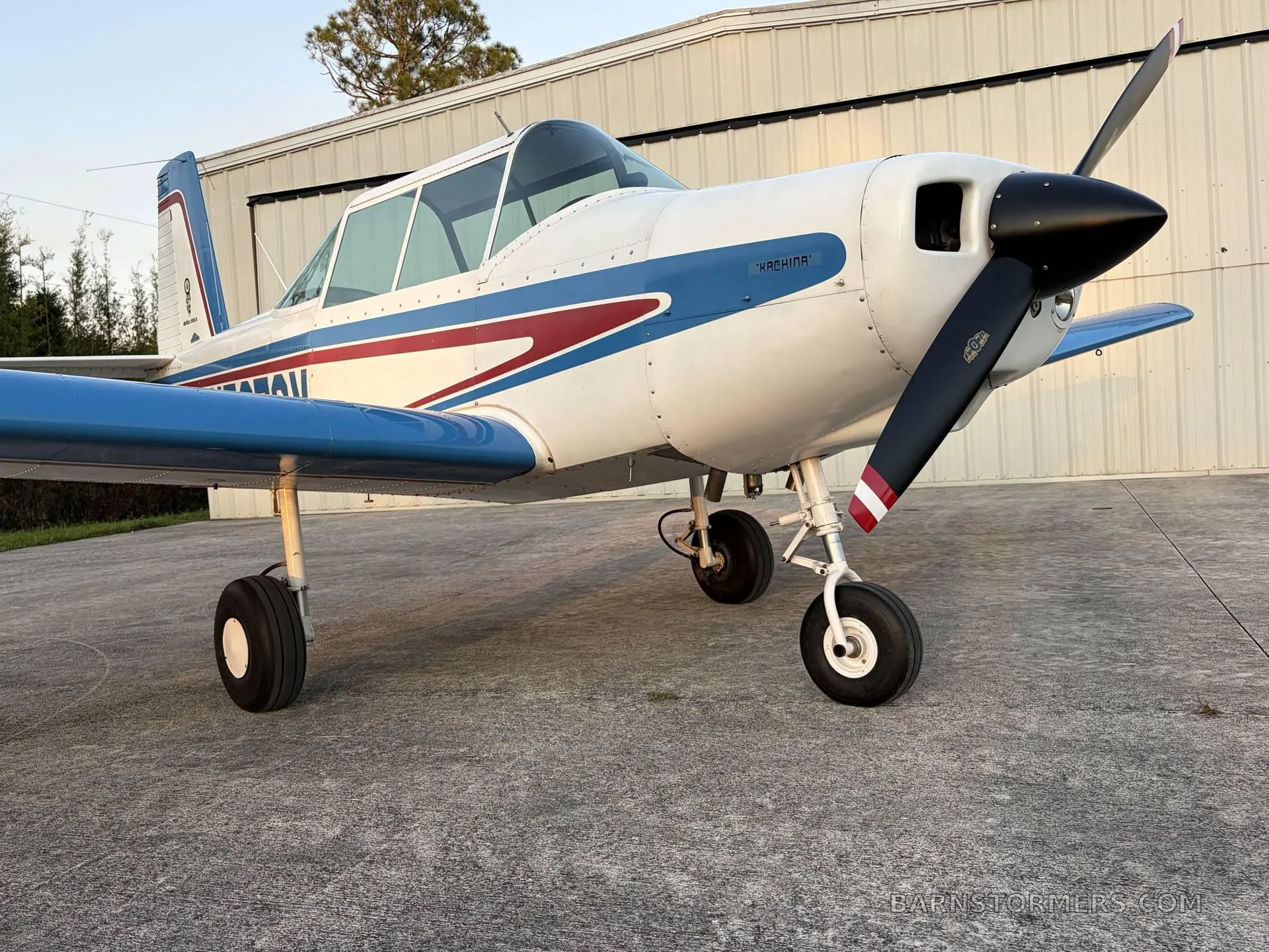 A white and blue single-engine aircraft with a red stripe, parked on a concrete pad in front of a hangar, viewed from the front-right.