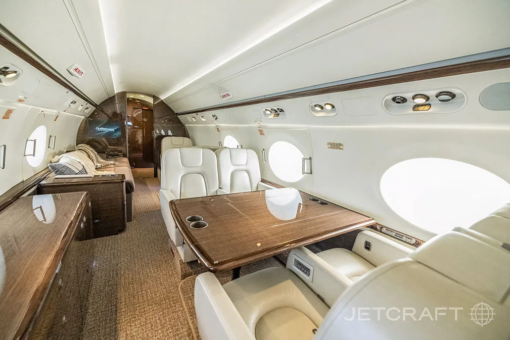 A private jet cabin section with a large wooden table, light leather seats, and a lounge area in the background.