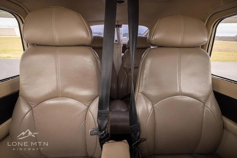 The tan leather front seats of a Cessna 182 Skylane cabin, with seatbelts fastened, viewed from the rear.