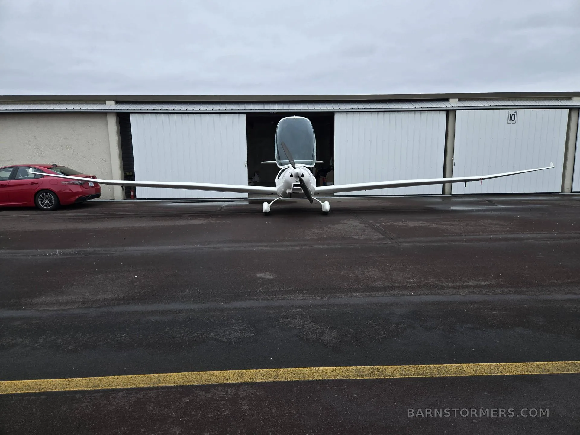 A white motor glider with its canopy open is parked on an apron in front of a hangar.