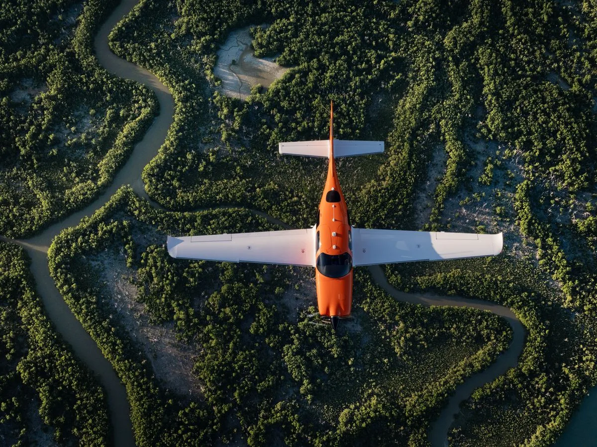 An orange and white single-engine aircraft flies over a winding river through lush green trees, viewed from directly above.