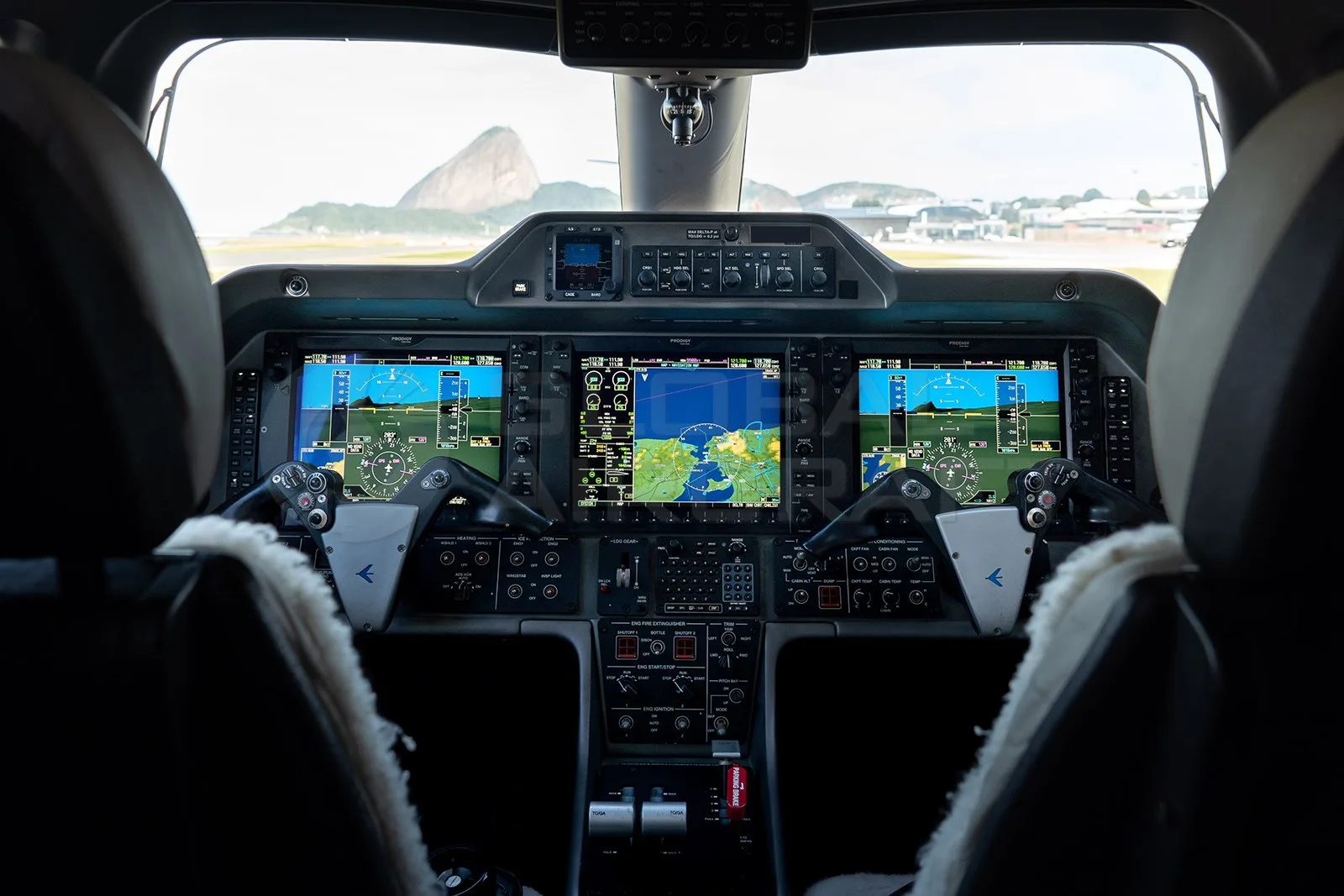 The cockpit of an Embraer Phenom 100 shows multiple display screens and controls, with a view of a runway and mountains through the windshield.