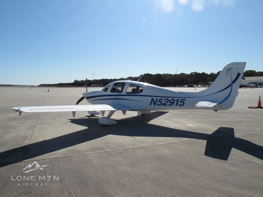 A white Cirrus SR20 aircraft with blue and grey stripes is parked on a concrete ramp, viewed from the right side.