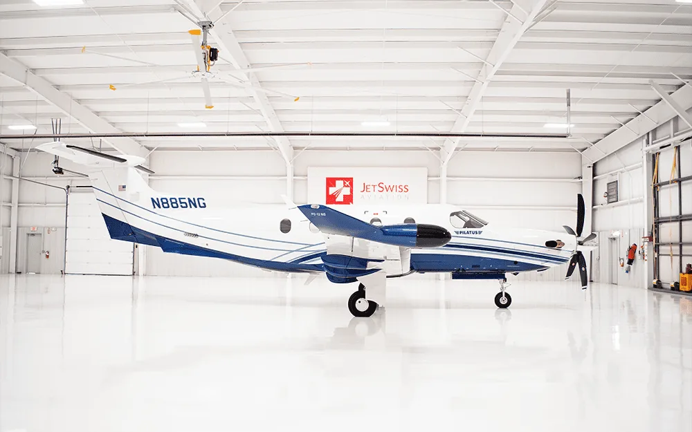 A white and blue Pilatus PC-12 aircraft, tail number N885NG, is parked in a brightly lit hangar, viewed from the side.