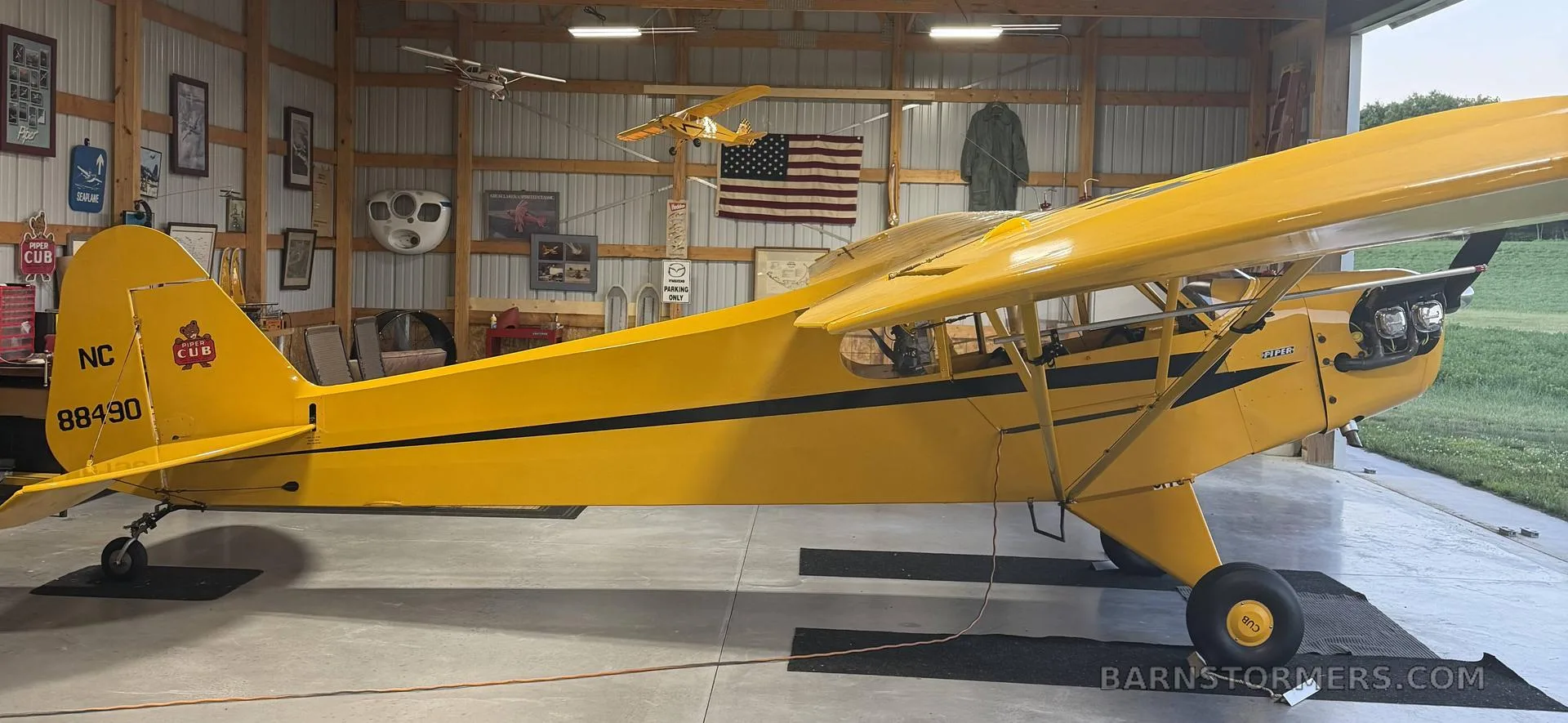 A side view of a yellow Piper Cub aircraft with black stripes parked inside a hangar, showing the tail number NC88490.