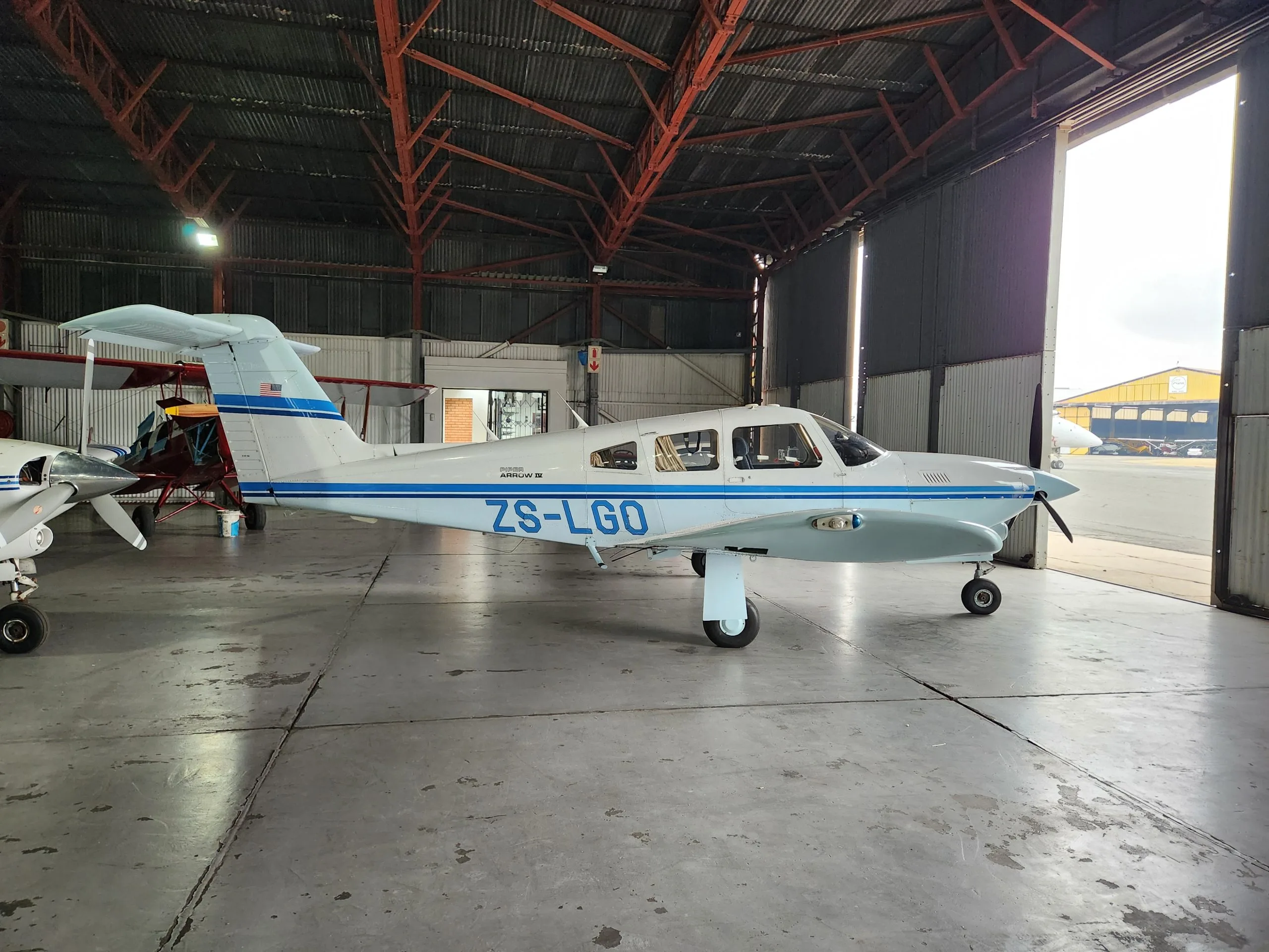 A white and blue Piper aircraft, registration ZS-LGO, is parked inside a hangar with the door open, viewed from the right side.