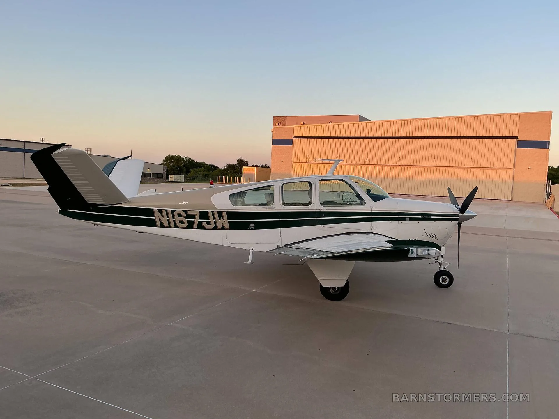 A side view of a white and dark green Beechcraft Bonanza (N167JW) parked on an airfield tarmac with hangars in the background.