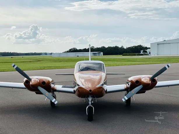 Head-on view of a copper and white Piper Comanche twin-engine aircraft with propellers visible.