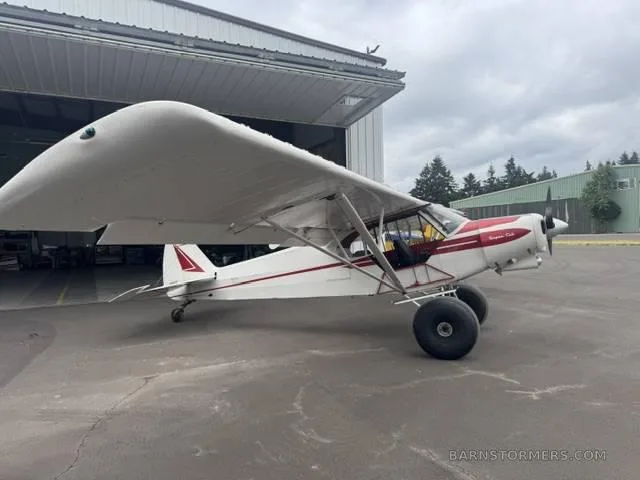 A white and red Super Cub aircraft with large tundra tires parked on an asphalt apron next to a hangar.