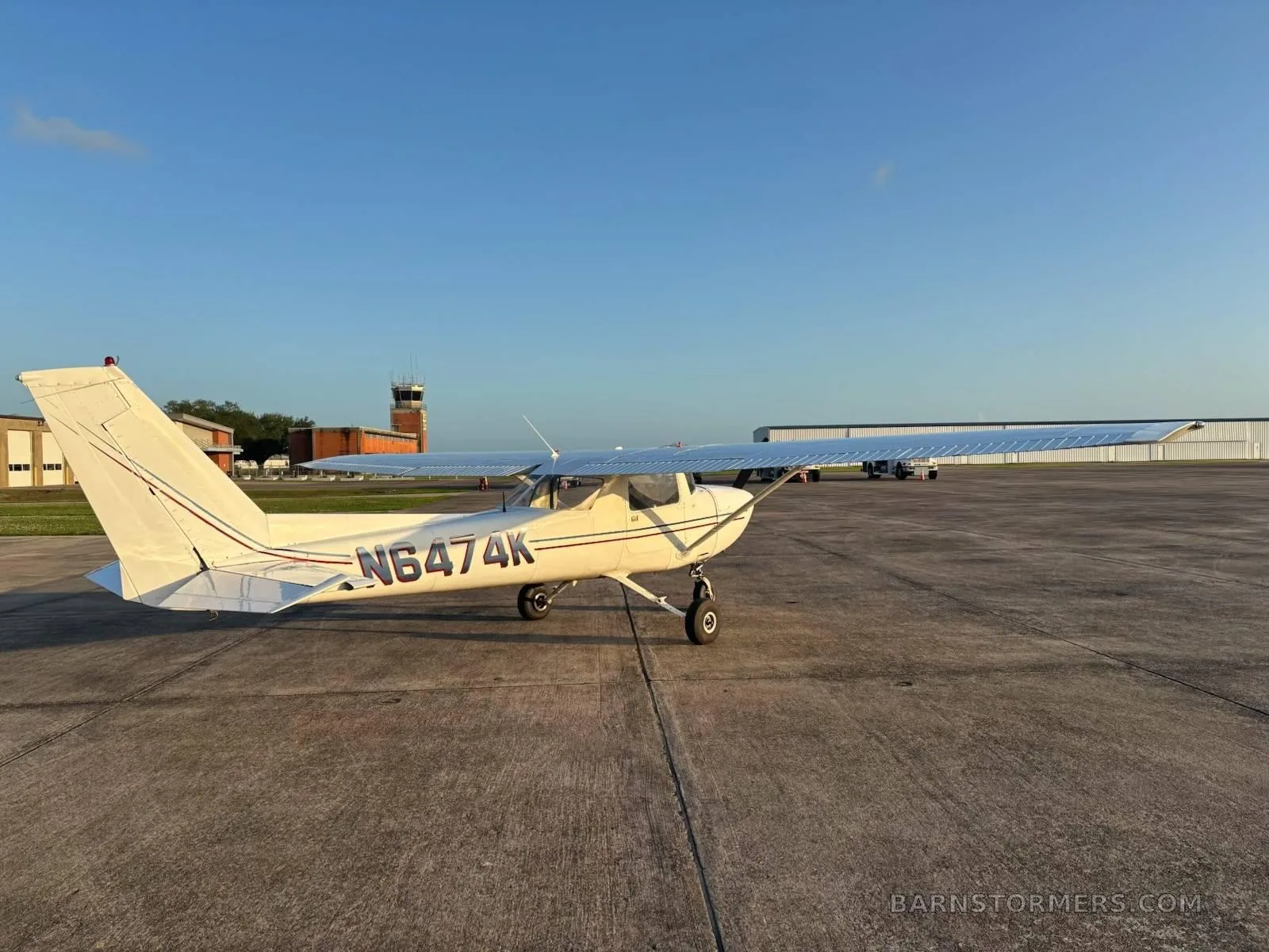 A white Cessna 172 (or similar) with tail number N6474K is parked on an airfield tarmac with a control tower in the background.