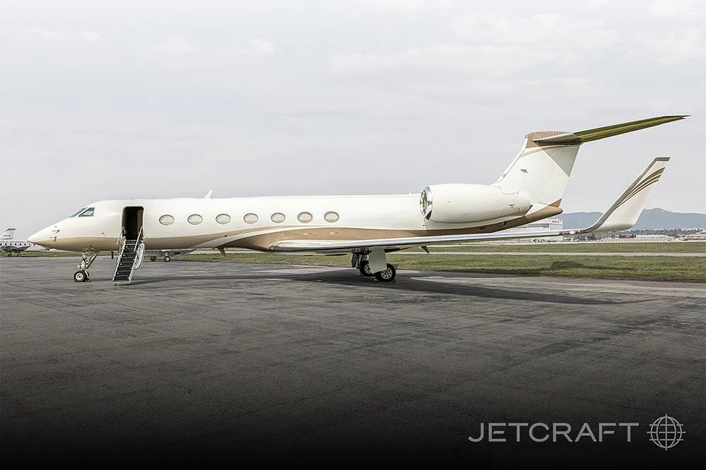A Gulfstream G650 business jet with a white and gold paint scheme is parked on the tarmac with its door open and stairs extended.